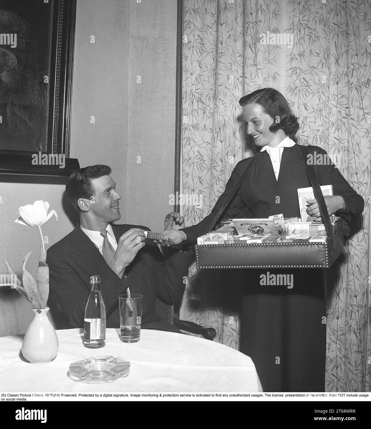 In the 1950s. A cigarette girl going around the tables in the restautrant selling cigarettes from her box seen in front of her. A man is seen recieving a package and has a coin in his hand to pay.  Sweden 1953  Kristoffersson ref  BL39-12 Stock Photo