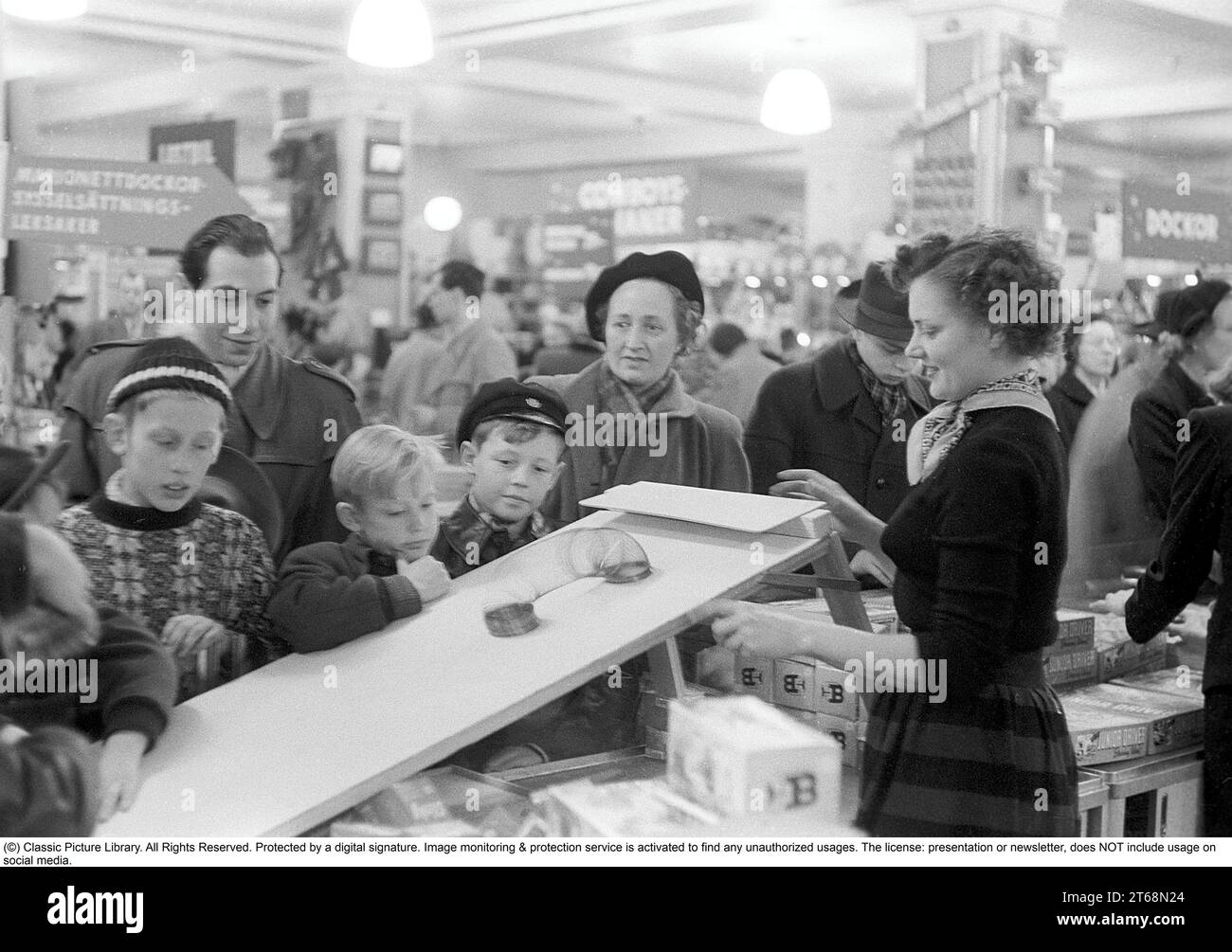 Toys in the 1950s. Interior from the NK department store in Stockholm. A clerk in the toy department shows how the Walking Spring works. A classic toy from the era. The spring was started, for example, on a staircase and then wandered down by itself. The children and adults gathered around the demonstration watch in fascination. Stockholm 1953. Kristoffersson ref 1-2 Stock Photo