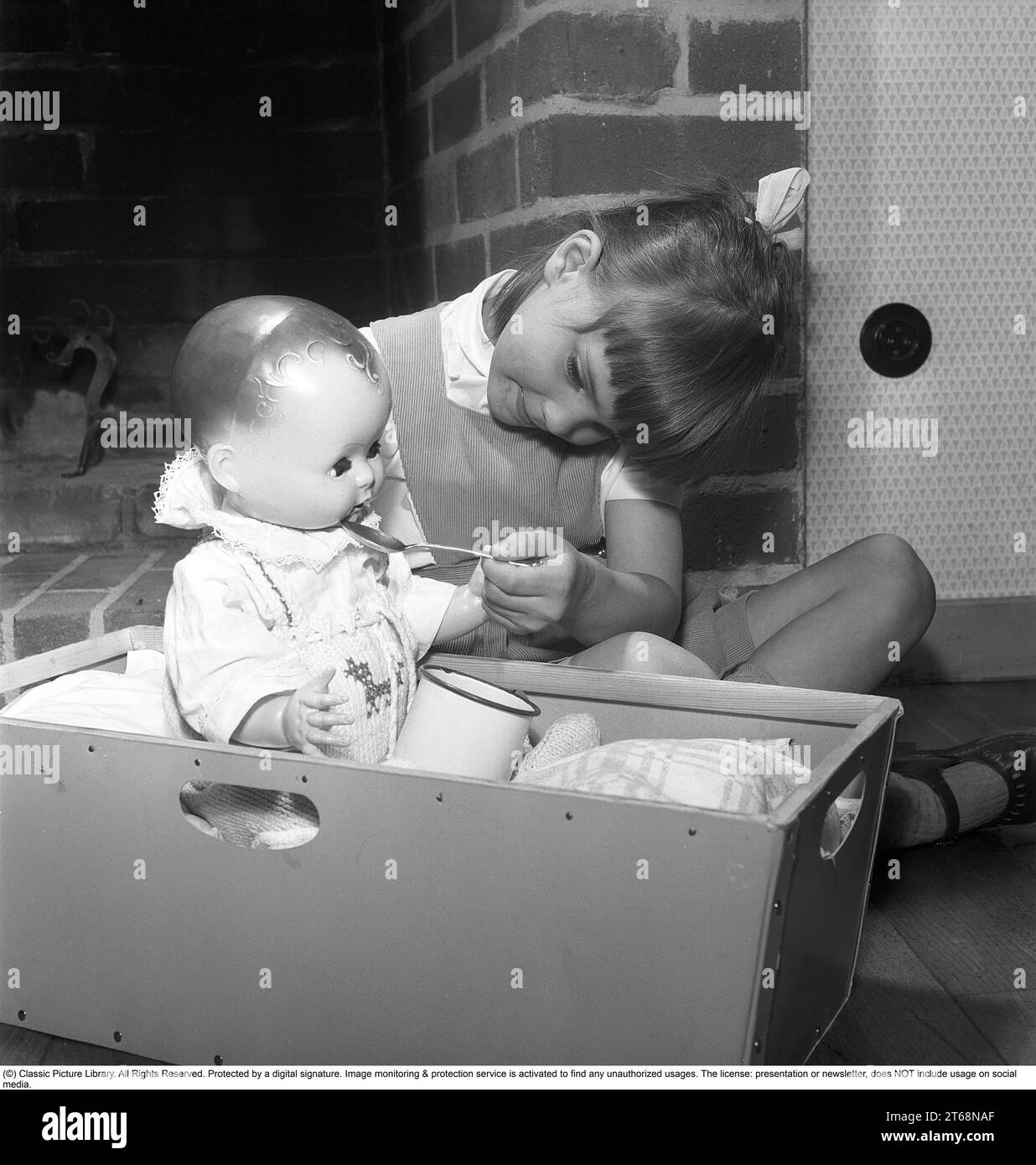 A girl with a doll in the 1950s. A little girl is playing with her doll. She puts a spoon to its mouth like you do when you give a child their medicine. Sweden 1956. Kristoffersson ref BS40-5 Stock Photo