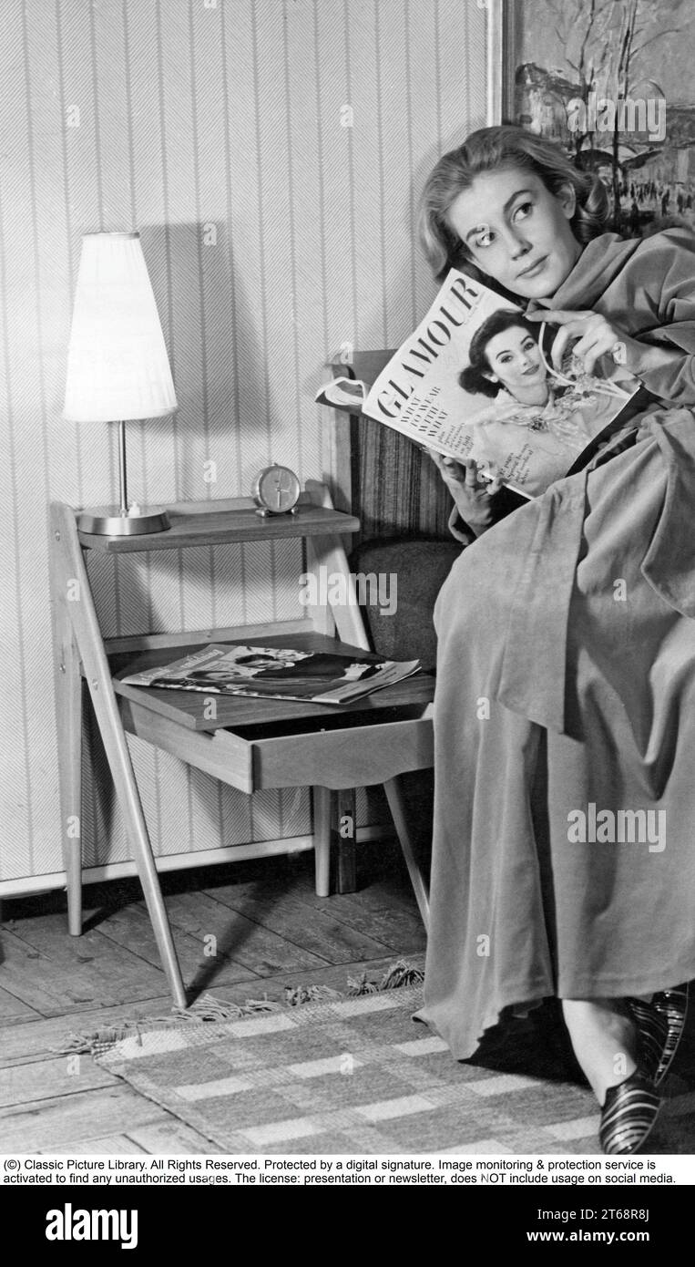 In the 1950s. A woman is seen reading a magazine somewhat sitting lying on a bed with a wooden sideboard visible. Sweden 1957 Stock Photo