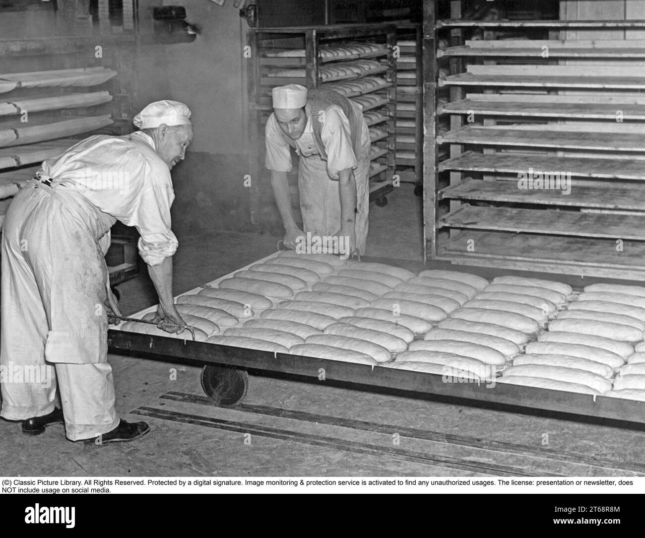 Bakery in the 1930s. Two men working in the bakery, handling the loafs of bread ready to be put into the oven. This is a industrial bakery, baking thousands of bread. Sweden 1939 Stock Photo