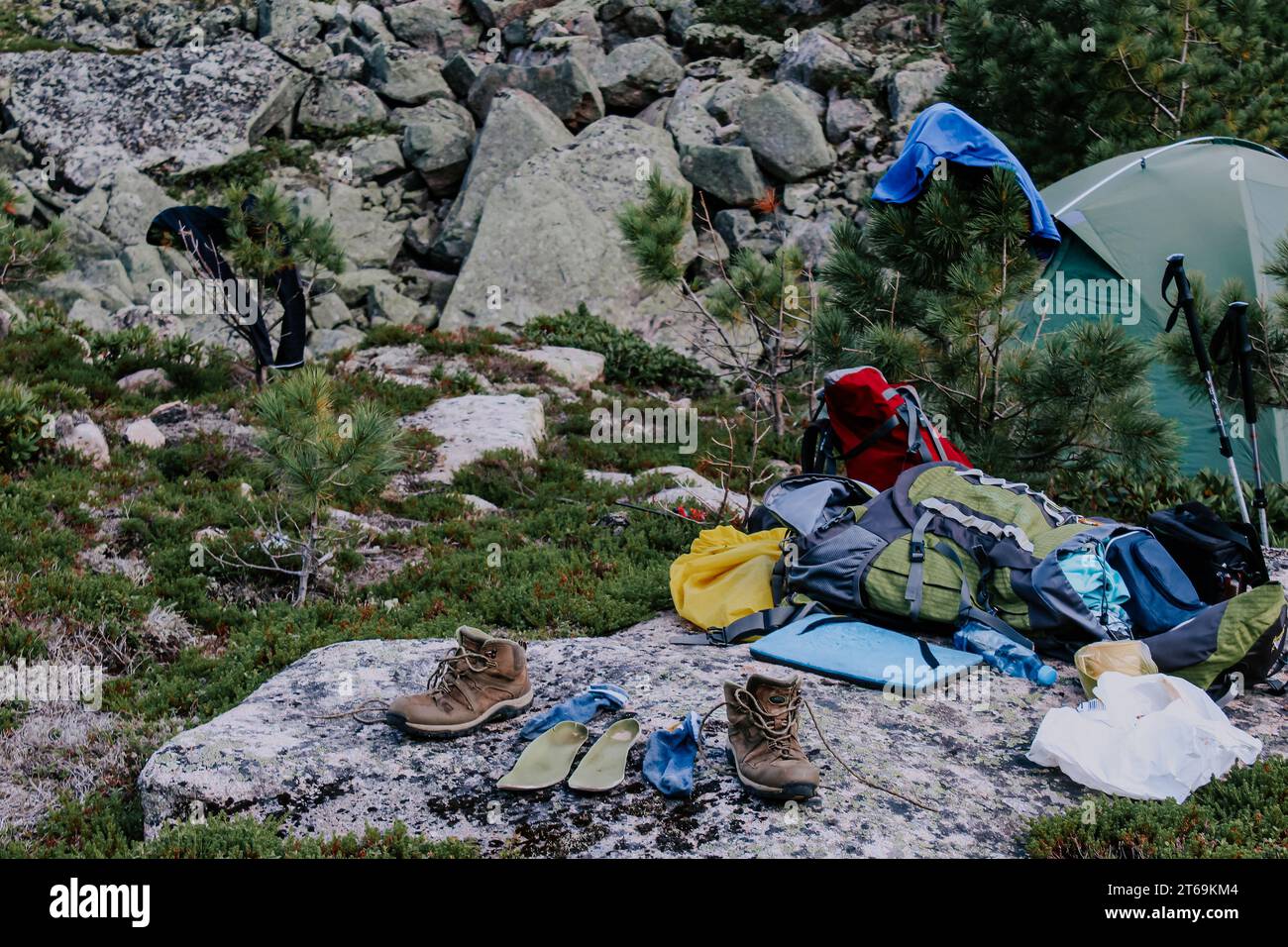 Equipment in the forest: tourist tent, clothes and shoes to dry on tree branches and stones at summer day. Tourist camping on a hike. Stock Photo