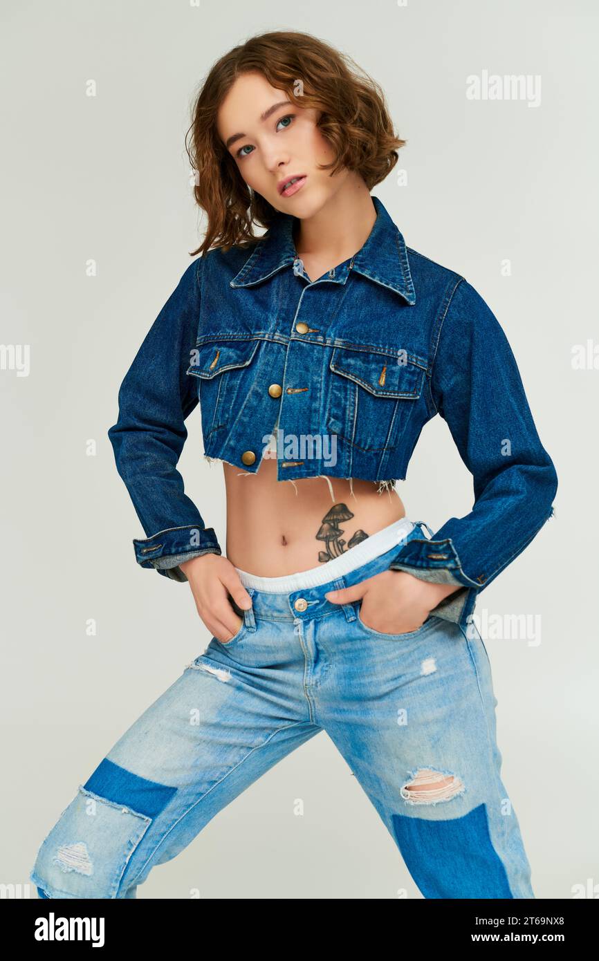 pretty model in trendy cropped denim jacket posing with hands in pockets of jeans on grey background Stock Photo