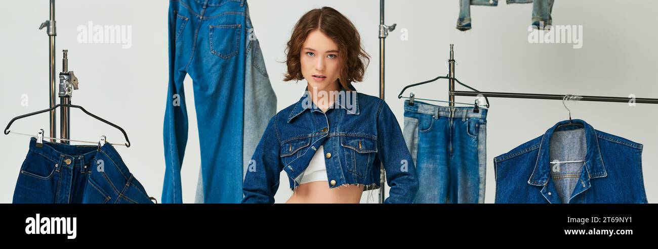 pretty model in cropped blue jacket posing among denim clothes on hangers on grey backdrop, banner Stock Photo