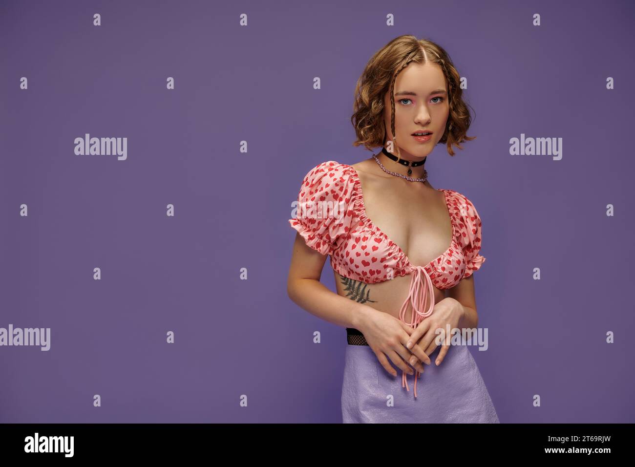 young gen z woman in cropped top with hearts pattern and skirt posing on purple background Stock Photo