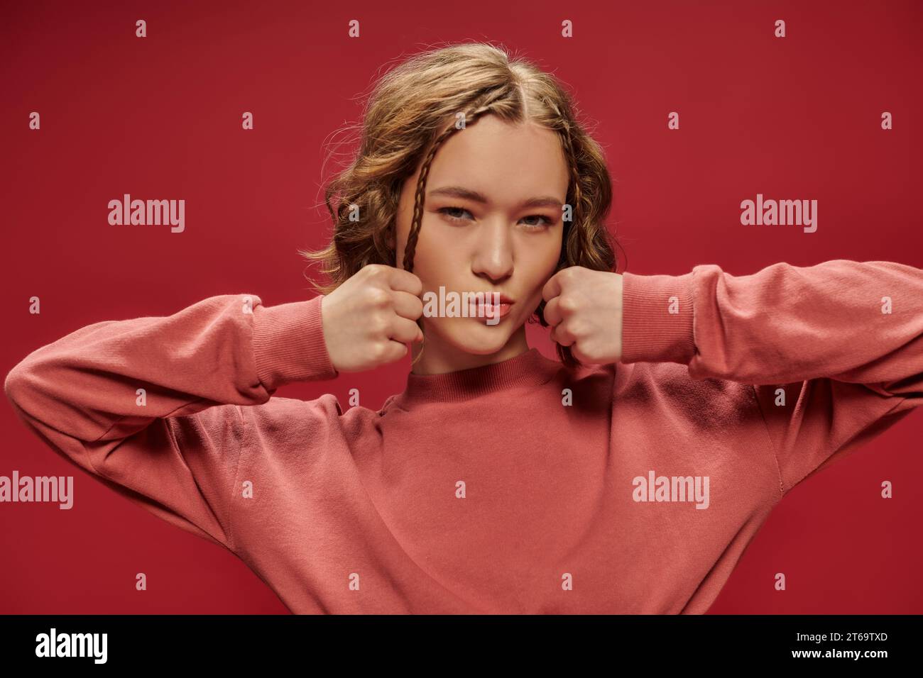 cute gen z girl with short hair and braids posing with clenched fists and looking at camera on red Stock Photo