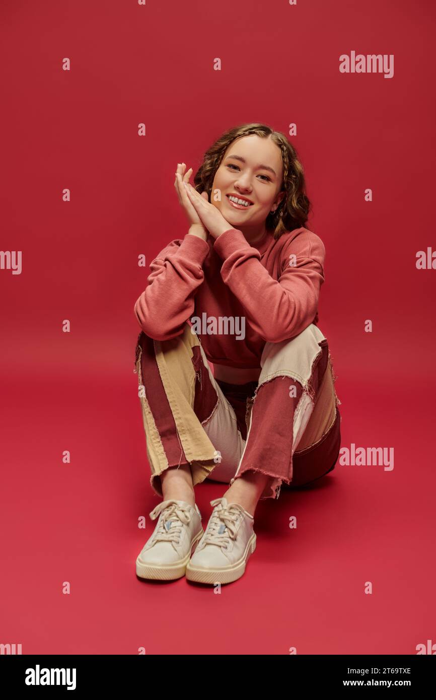 joyful woman in patchwork pants and cropped long sleeve sitting on red backdrop, youth culture Stock Photo