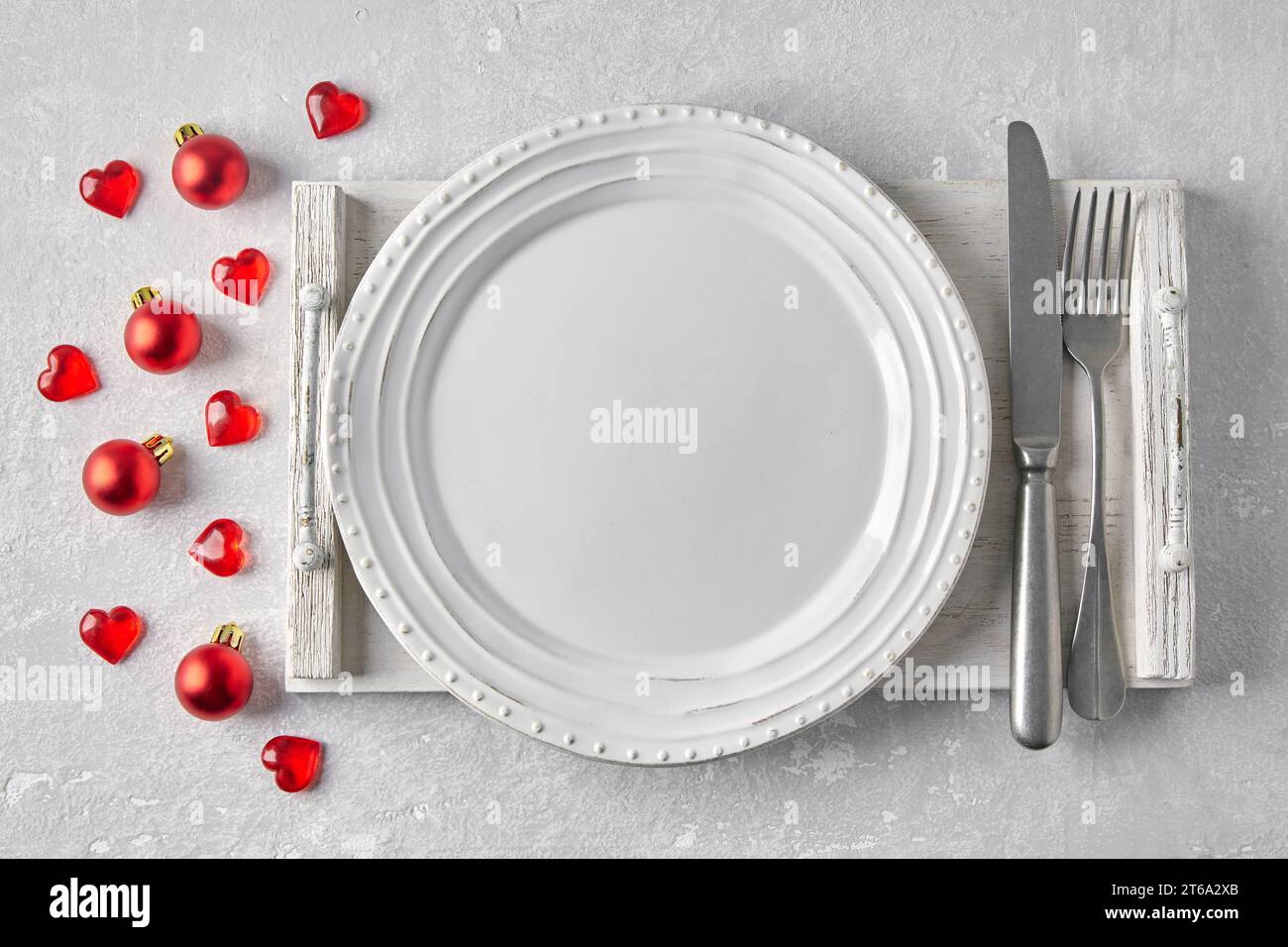 Empty white plate with cutlery on a white wooden tray on a gray concrete table surrounded by red Christmas balls and hearts. Template for presenting f Stock Photo