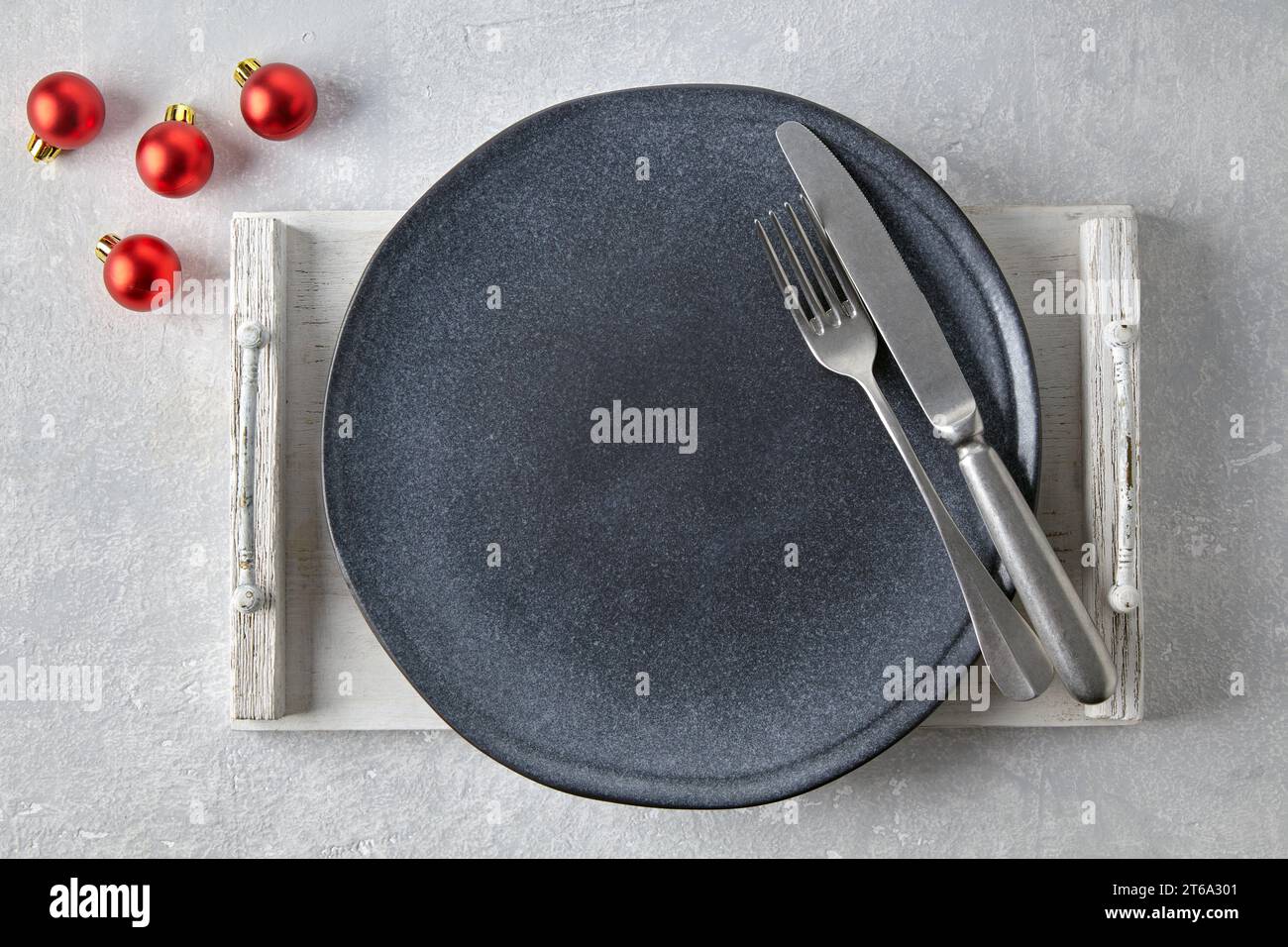 Empty dark gray plate with cutlery on a brown wooden tray on a gray concrete table surrounded by red Christmas balls. Template for presenting food in Stock Photo