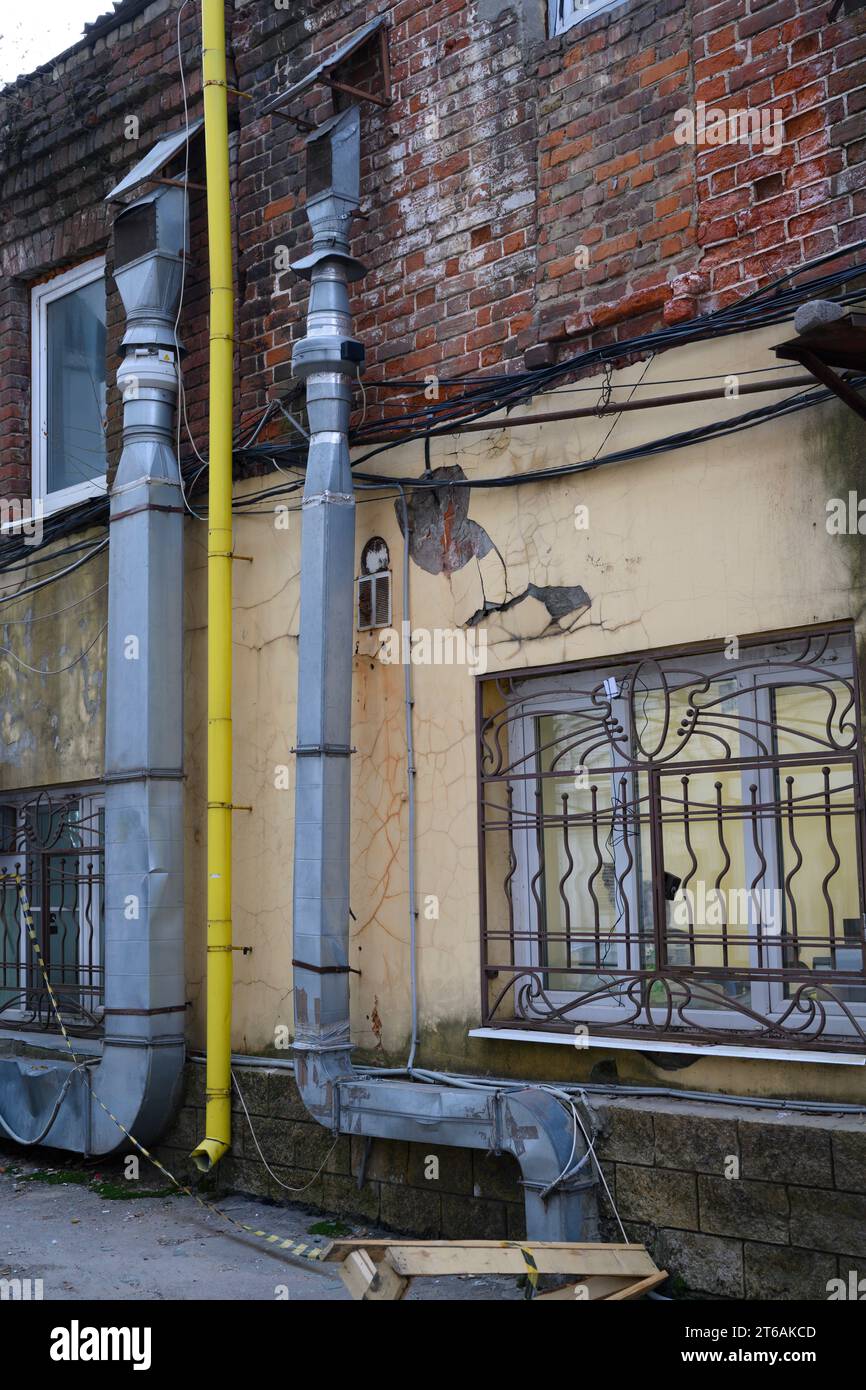 Urban landscape with a brick peeling wall with barred windows, ventilation ducts and a yellow downpipe Stock Photo