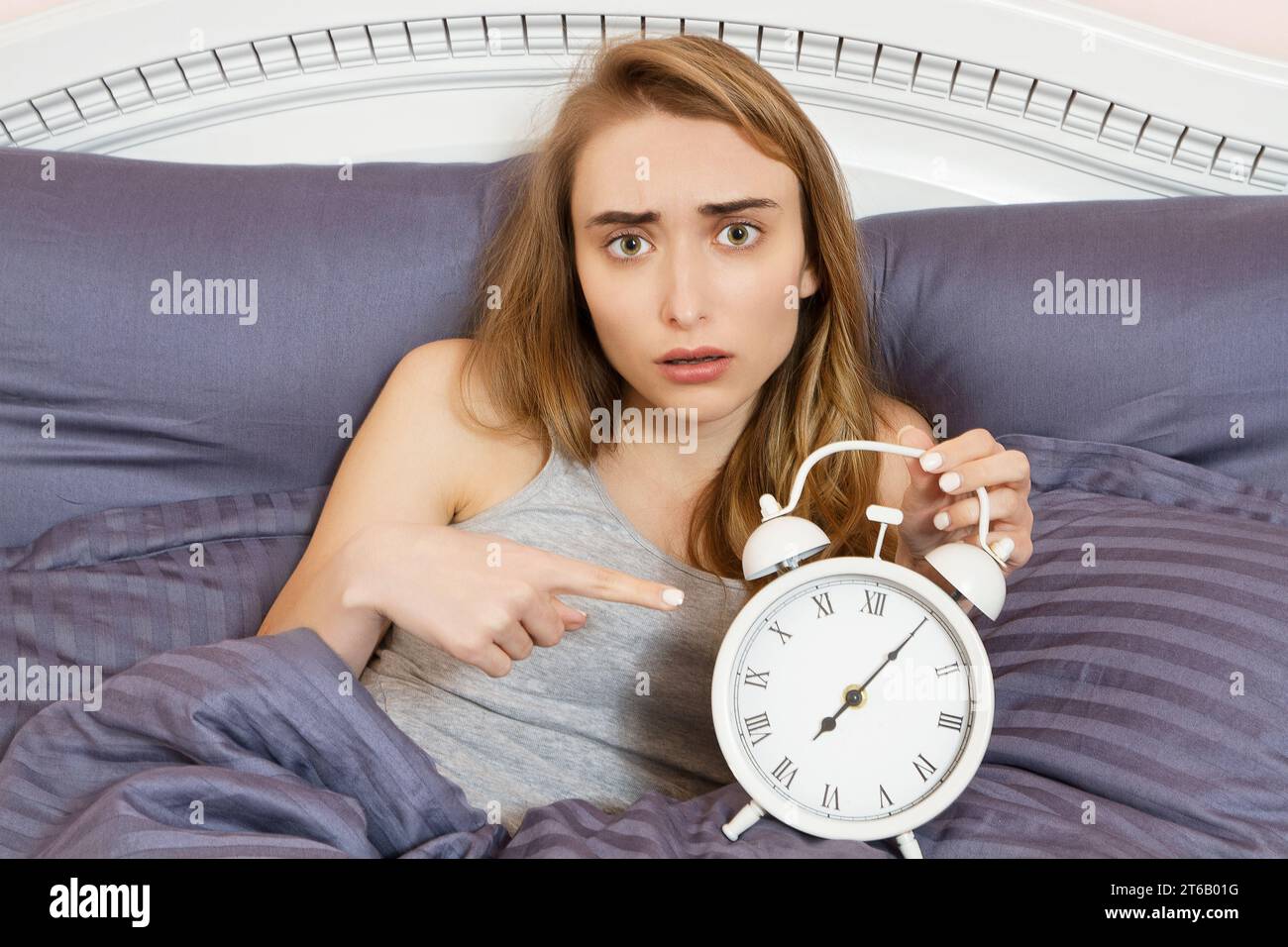 Shocked young woman waking up with alarm, girl point on clocks in bed