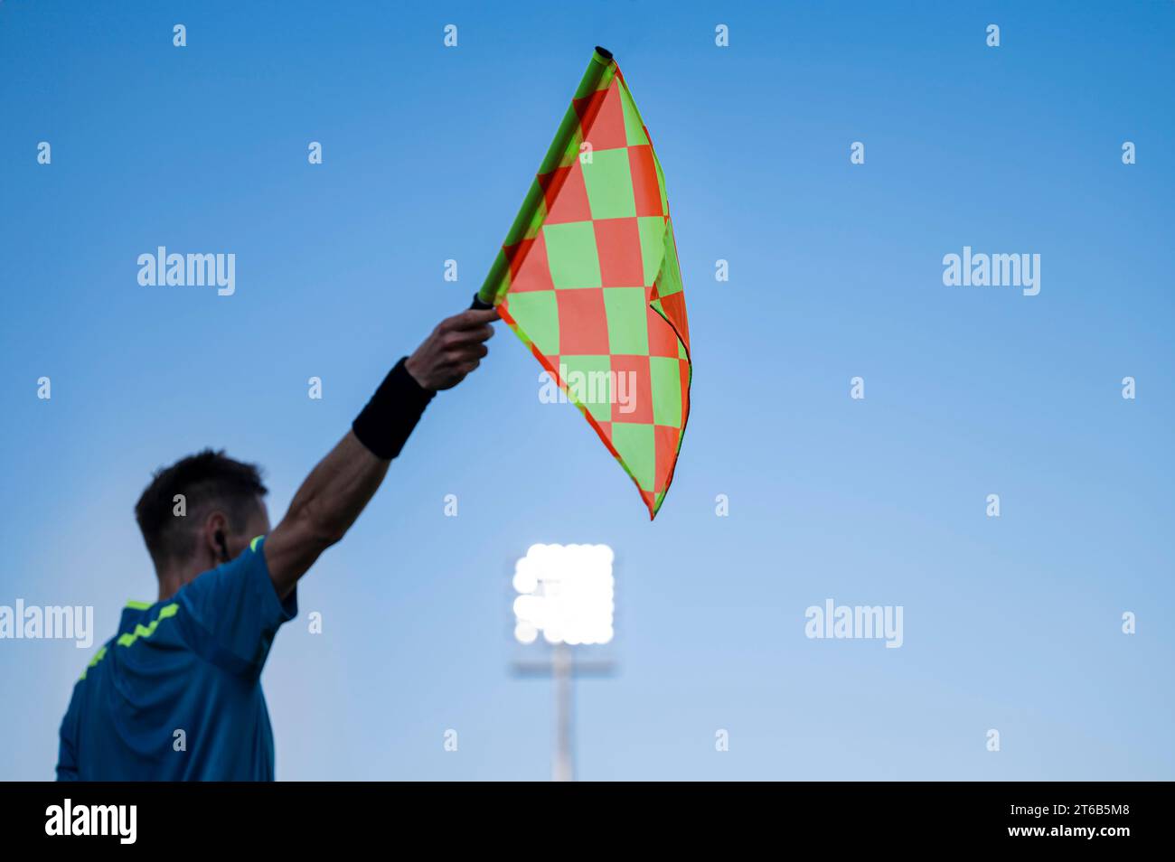 Raised flag of the sideline referee during football match, blue sky and lamps in the background. Stock Photo