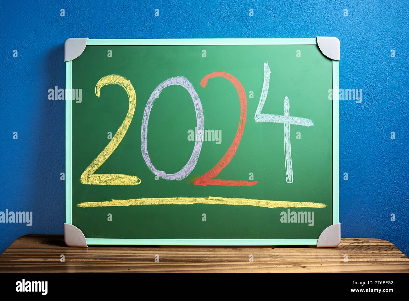 Front view of the number 2024 written in chalk on green chalkboard placed on wooden table against flat blue wall. New year and back to school concept. Stock Photo