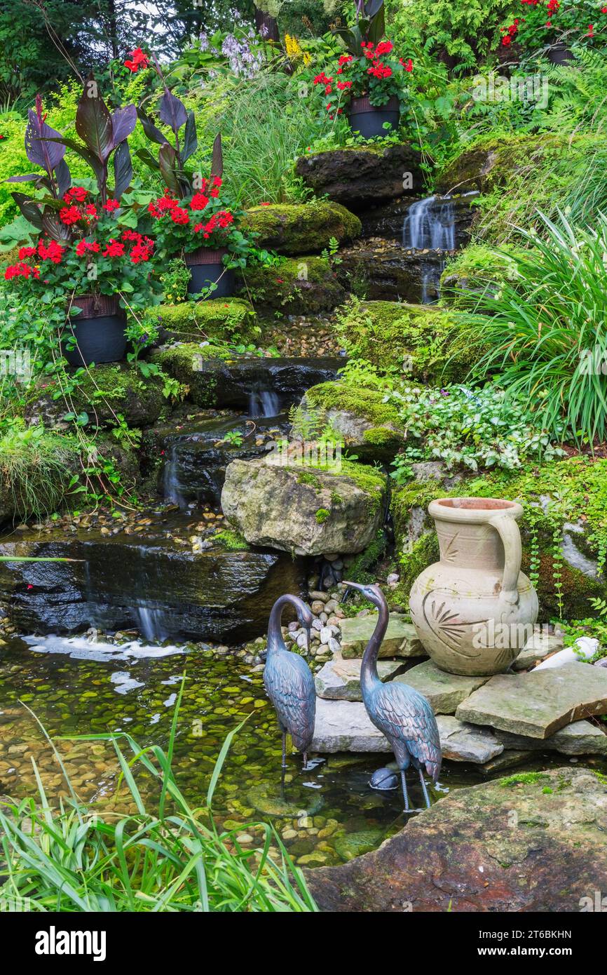 Aquatic bird sculptures and ceramic jug in pond with waterfall bordered by Lysimachia nummularia 'Aurea' - Golden Creeping Jenny, Lamium - Deadnettle. Stock Photo