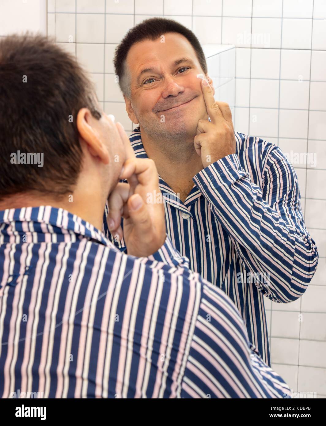 A man with a face of different ages applies cream in front of the bathroom mirror Stock Photo
