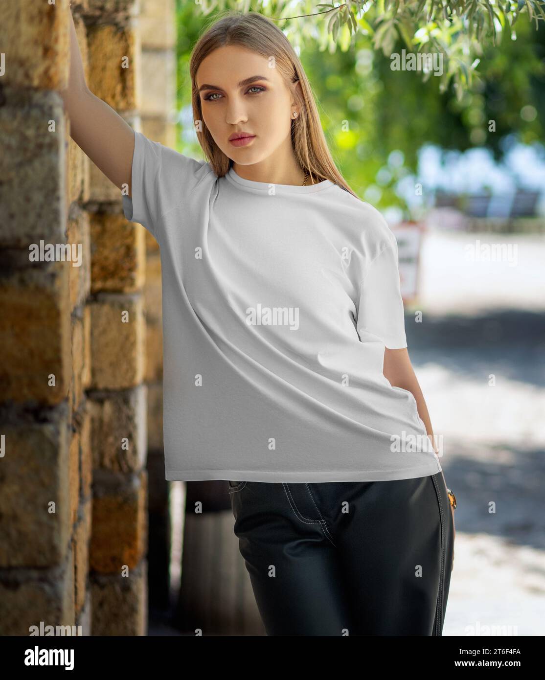 Mockup of a white T-shirt on a girl, leaning on a brick wall, on the background of trees, front view. Women's shirt template, presentation of clothes Stock Photo