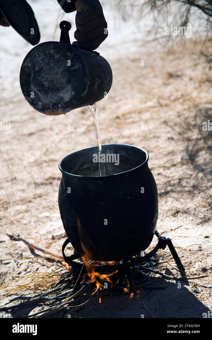 A man (camel trekking guide Nasser) pours water into a large pot over a fire in the desert. Stock Photo
