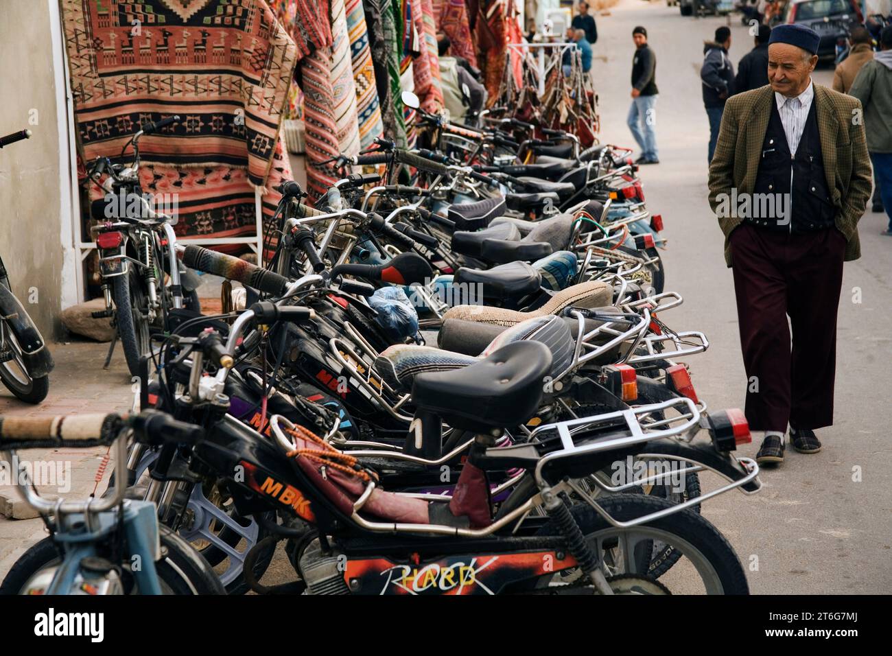 An old man wearing a fez walks past a row of motorcycles (mopeds). Stock Photo
