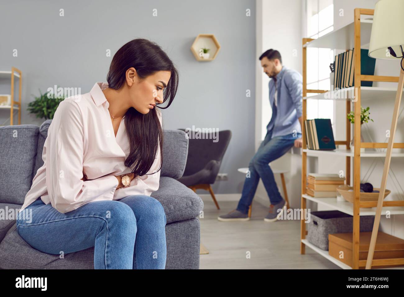 Upset young woman sitting on sofa and a man in background at home. Quarrel and divorce concept. Stock Photo
