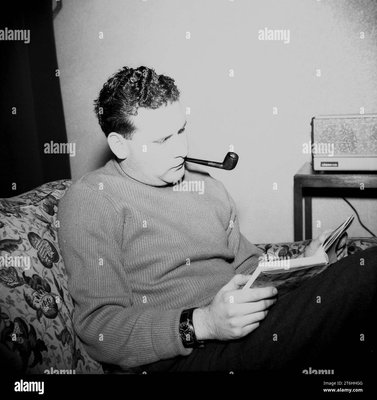 1960s, historical, a man relaxing, sitting in a flower patterned lounge chair smoking a pipe, reading a book, radio of the era beside him, England, UK. Stock Photo