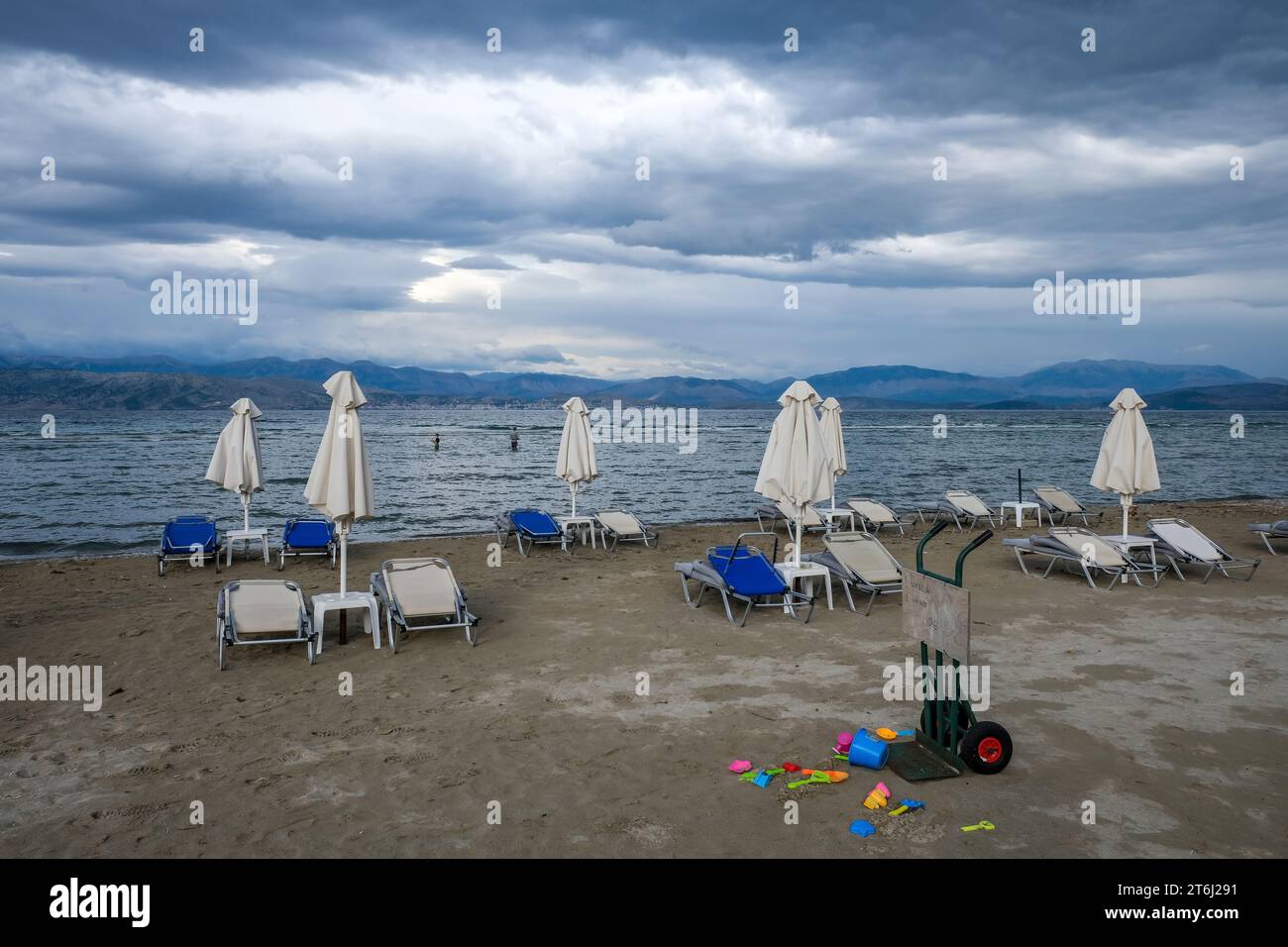 Kalamaki, Corfu, Greece, sun beds and umbrellas on Kalamaki beach in the northeast of the Greek island of Corfu, in the background the mainland of Albania with the seaside resort of Saranda. In front forgotten children's toys, bucket and shovel. Price board umbrella and sun lounger rental. Stock Photo
