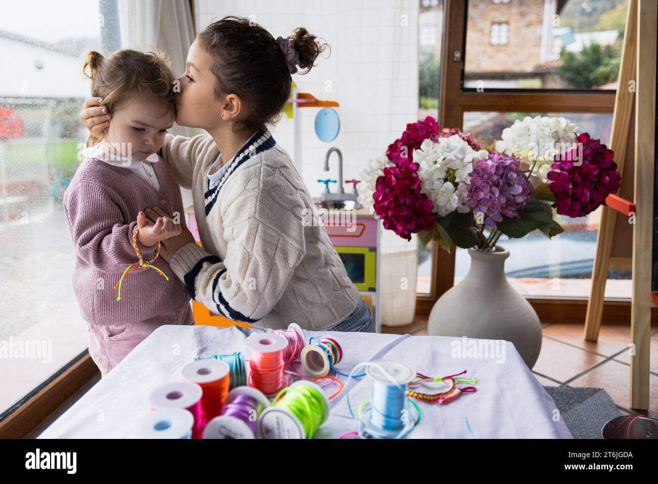 Two little girls sitting in the living room playing with colorful threads, older one kissing her sister Stock Photo
