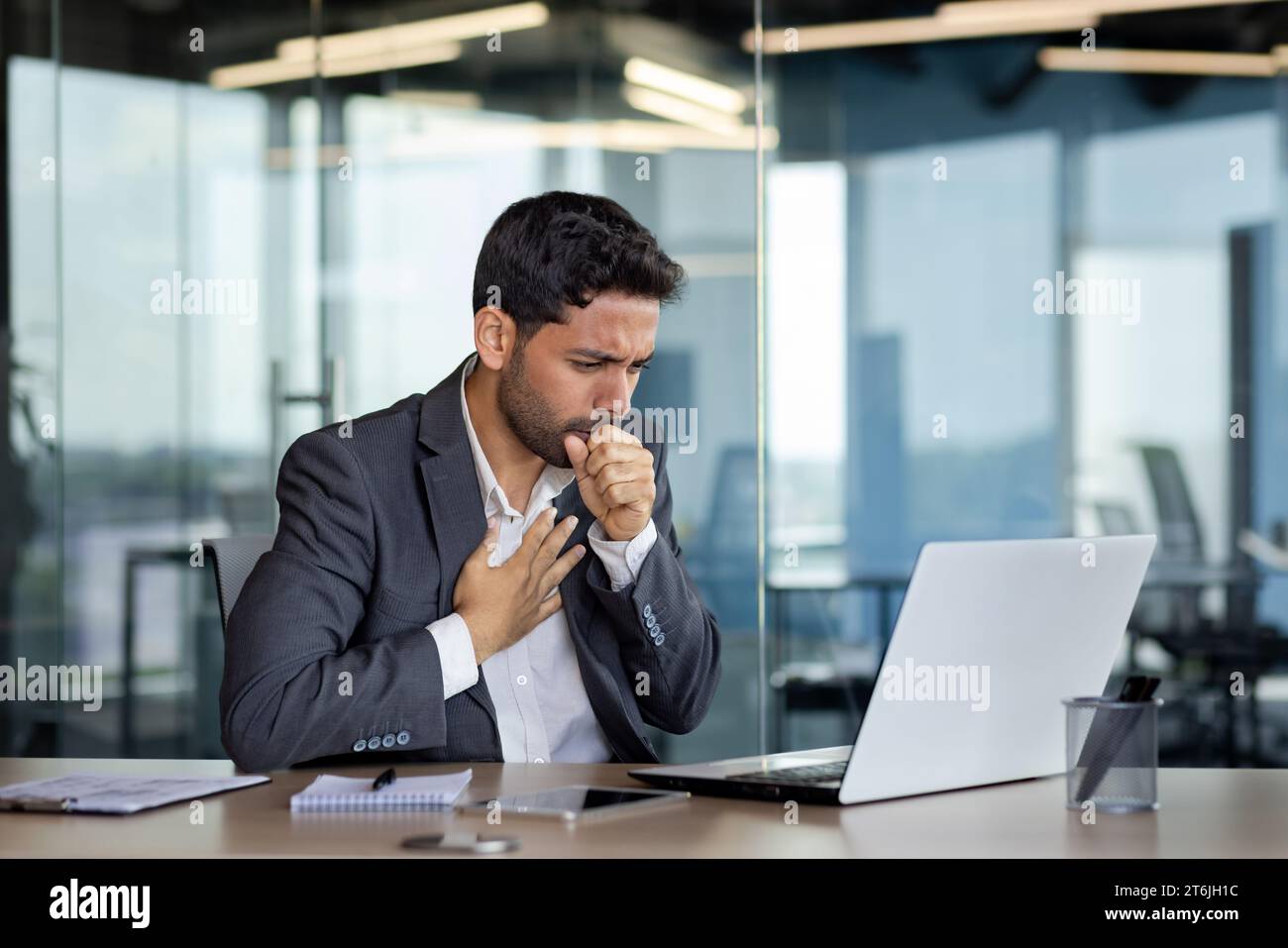 Sick businessman coughing inside office at workplace, man in business suit working with laptop