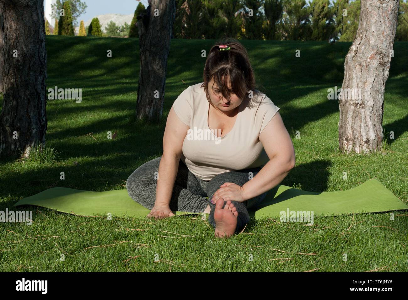 Overweight middle-aged woman stretching sitting on yoga mat in city park in summer. fat woman doing fitness to lose weight. Outdoor workout, yoga, str Stock Photo