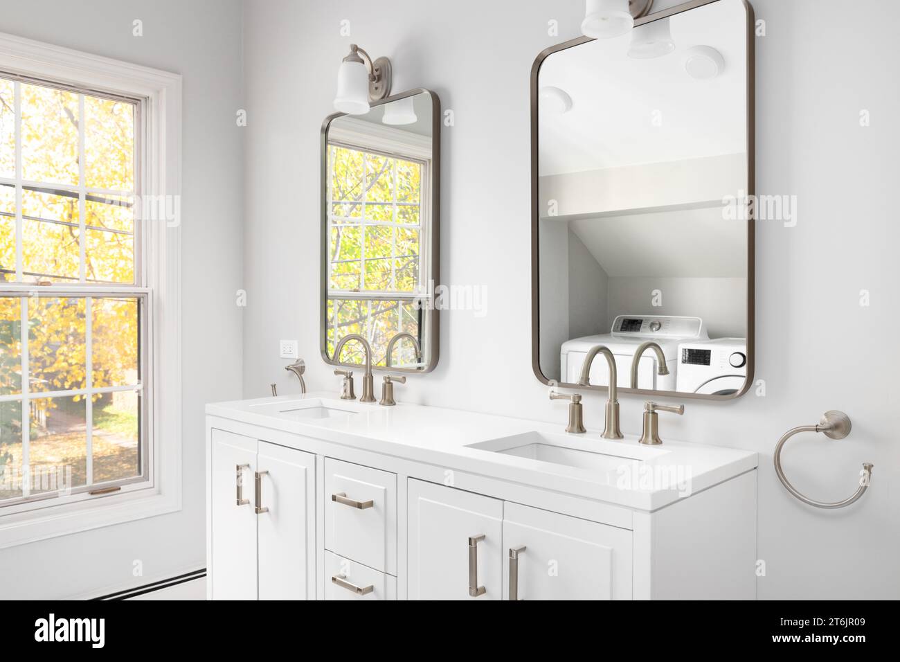A bathroom detail with a white cabinet, bronze faucets and mirrors, a view of the washer dryer in the reflection, and fall colors outside. Stock Photo
