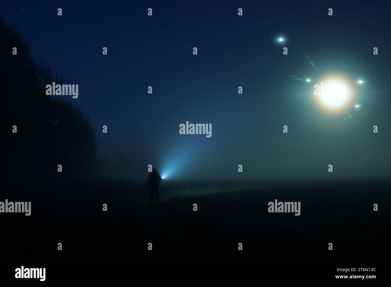 A man with a torch standing in the countryside on a misty summer night. With glowing UFO lights floating in the sky. Stock Photo