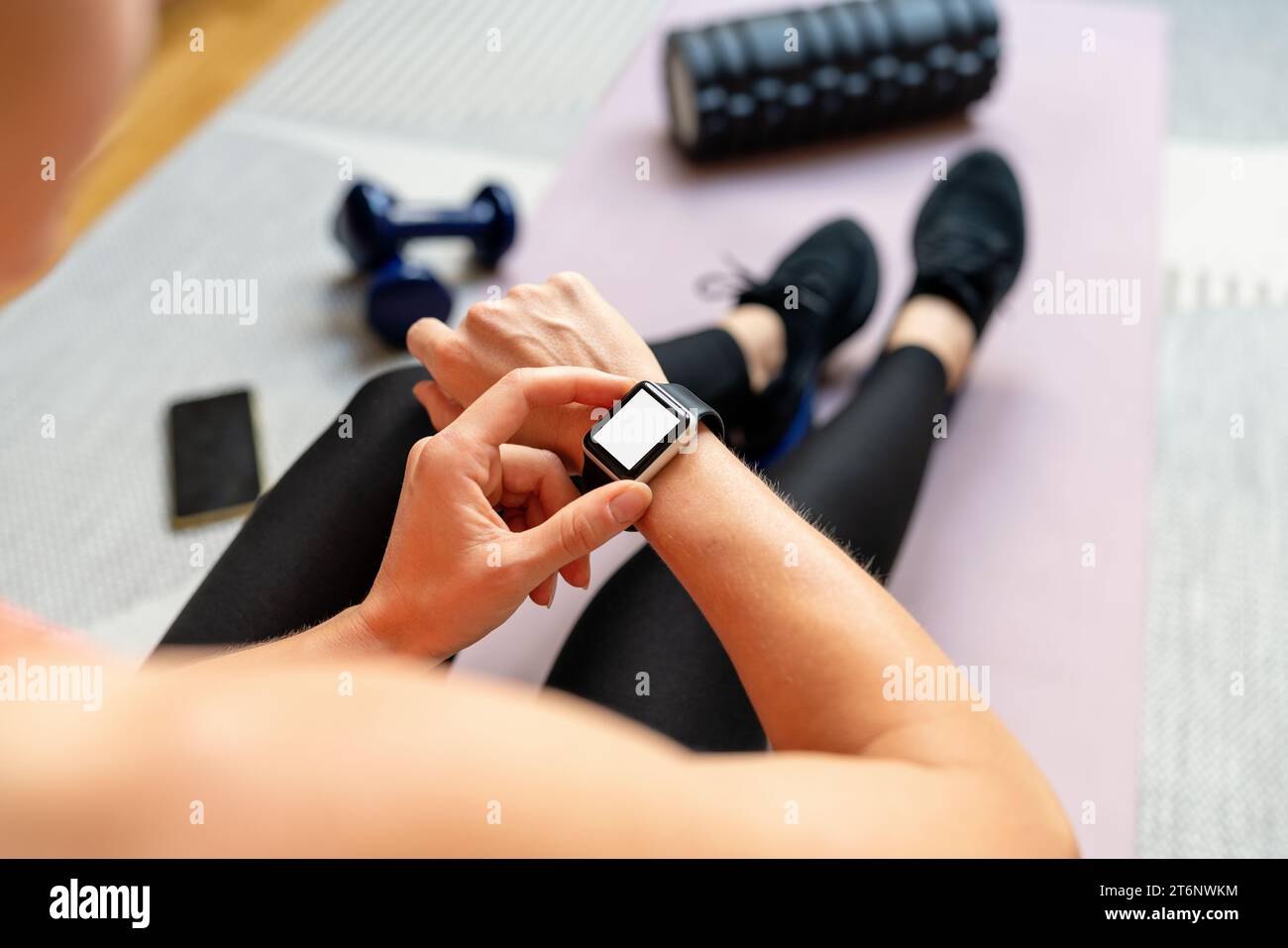 Over the shoulder view of female athlete looking at her smart digital watch with white blank screen while sitting on exercise mat. Stock Photo
