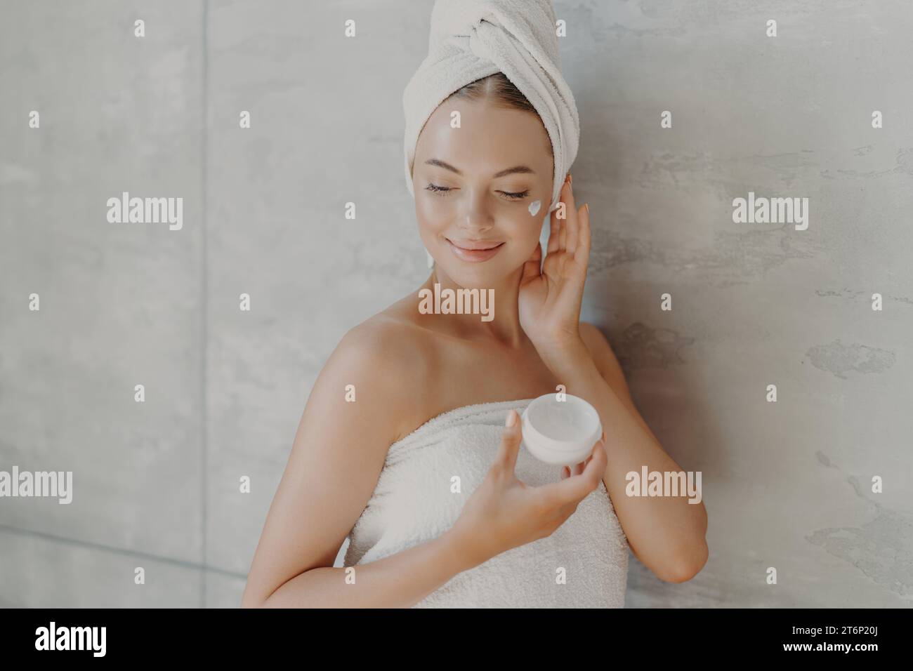 A woman with a towel turban applies cream, embodying a peaceful skincare moment Stock Photo