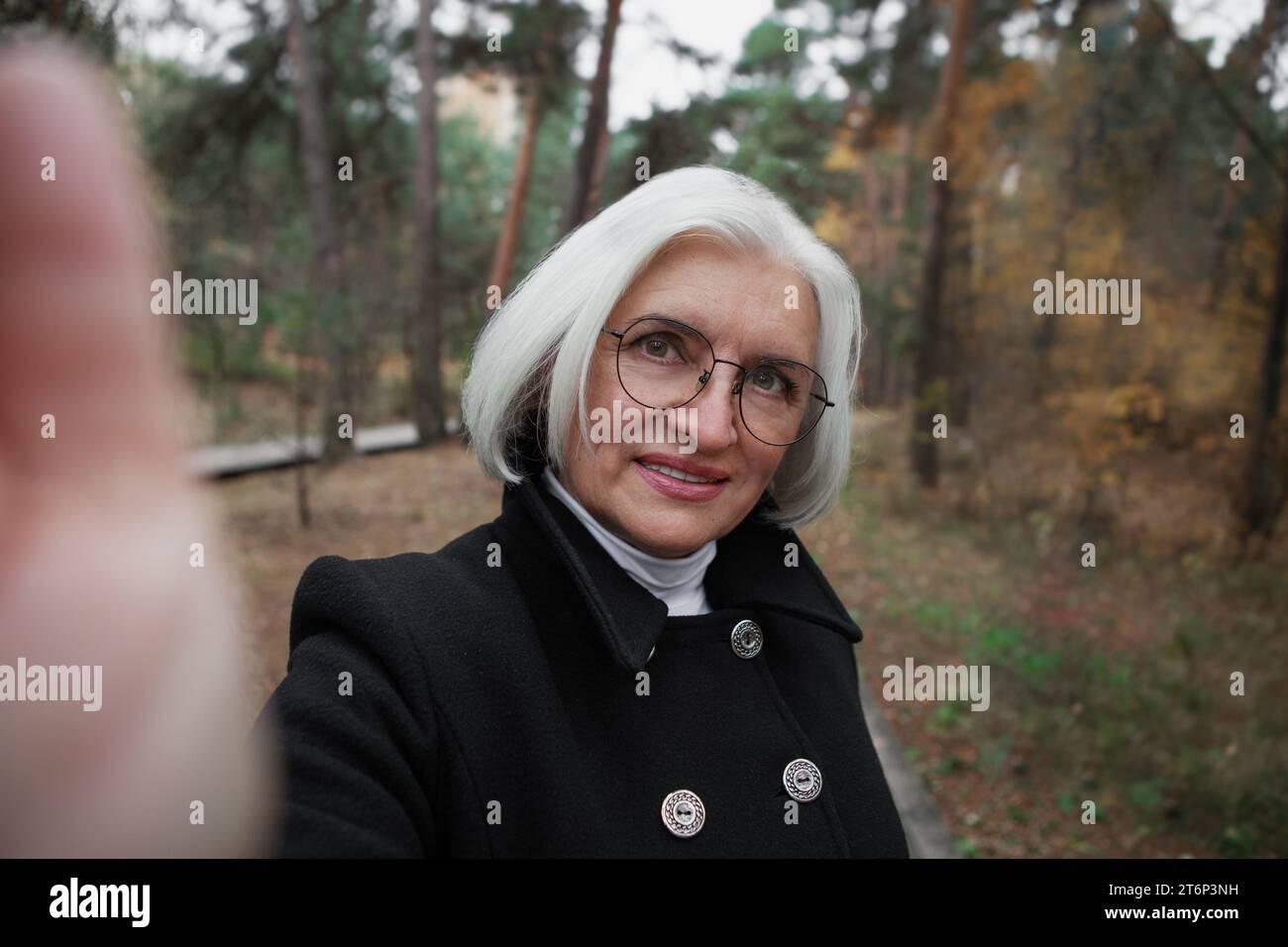 Authentic selfie portrait of attractive elderly 60 year old woman with gray hair and eyeglasses in nature forest or park. Real person, neutral color. Stock Photo