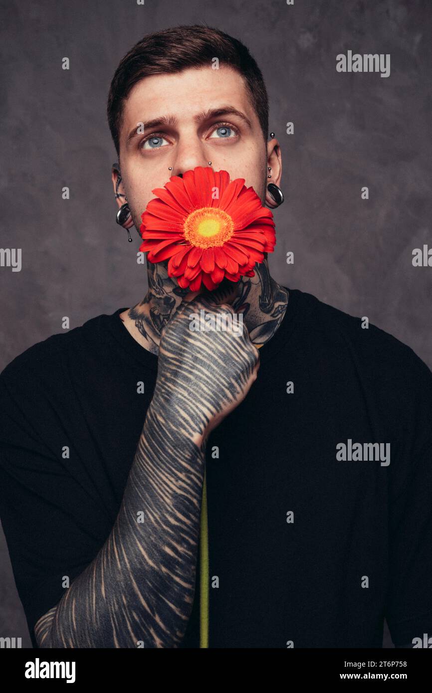 Contemplated young man with pierced nose ears holding red gerbera flower front his mouth against grey background Stock Photo
