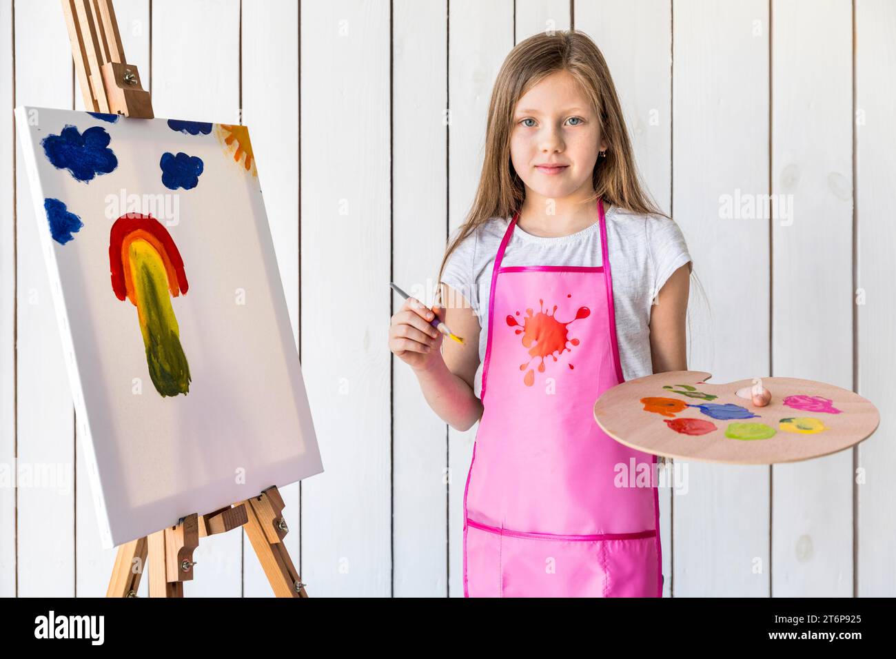 Portrait female artist holding paint brush palette standing against white wooden wall Stock Photo