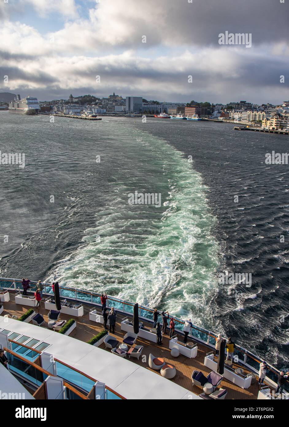 La ciudad de Alesund desde la salida de un crucero, estela de barco y cielo tormentoso. Turistas en la cubierta del barco tomando fotografías. Noruega Stock Photo