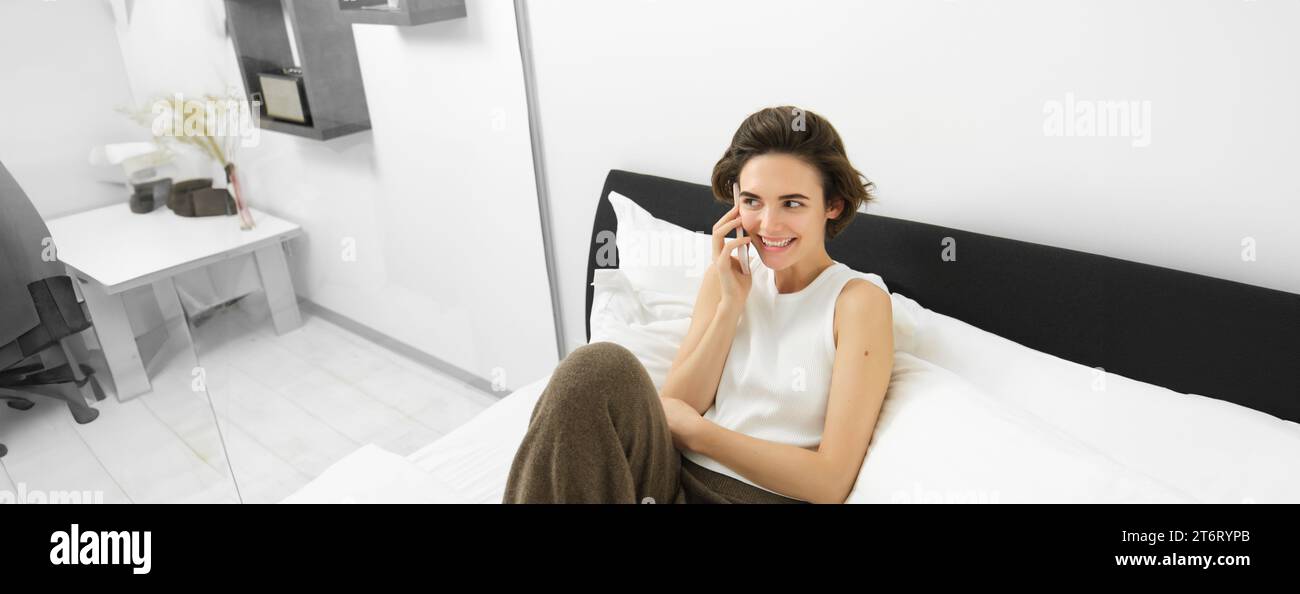 Chatty young woman laughing, talking on phone, calling friend, lying in bed and having conversation with someone, resting in bedroom Stock Photo