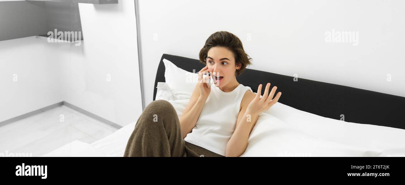 Chatty young woman laughing, talking on phone, calling friend, lying in bed and having conversation with someone, resting in bedroom Stock Photo