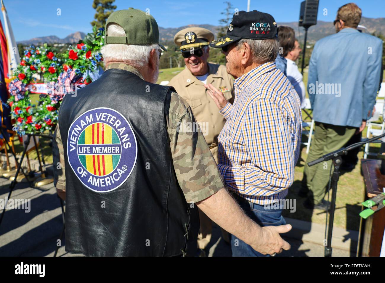 November 11, 2023, Santa Barbara, California, U.S: A Vet in a Vietnam Veterans of America Life Member vest and army shirt talks to other veterans, at the end of the annual Veteranâ€™s Day Ceremony at Santa Barbara Cemetery, â€œHonoring all who have servedâ€, hosted by Pierre Claeyssenâ€™s Veterans Foundation. A beautiful green wreath with red roses is in the background. (Credit Image: © Amy Katz/ZUMA Press Wire) EDITORIAL USAGE ONLY! Not for Commercial USAGE! Stock Photo