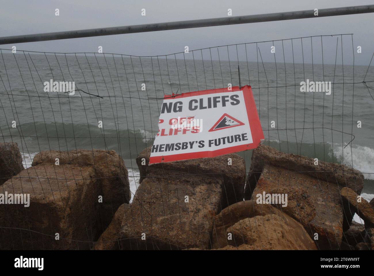 Ripped sign hanging on fence, asking beach visitors to avoid crumbling cliffs to protect Hemsby's future, blown by wind, during high seas. Hemsby Gap Stock Photo