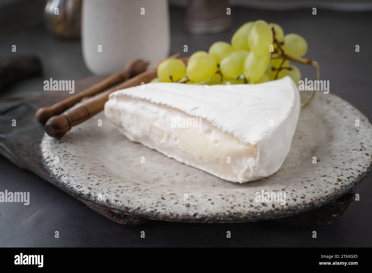 Soft brie camembert cheese with sweet grapes on a handmade clay plate. Side view of a piece of soft cheese with mold. Stock Photo