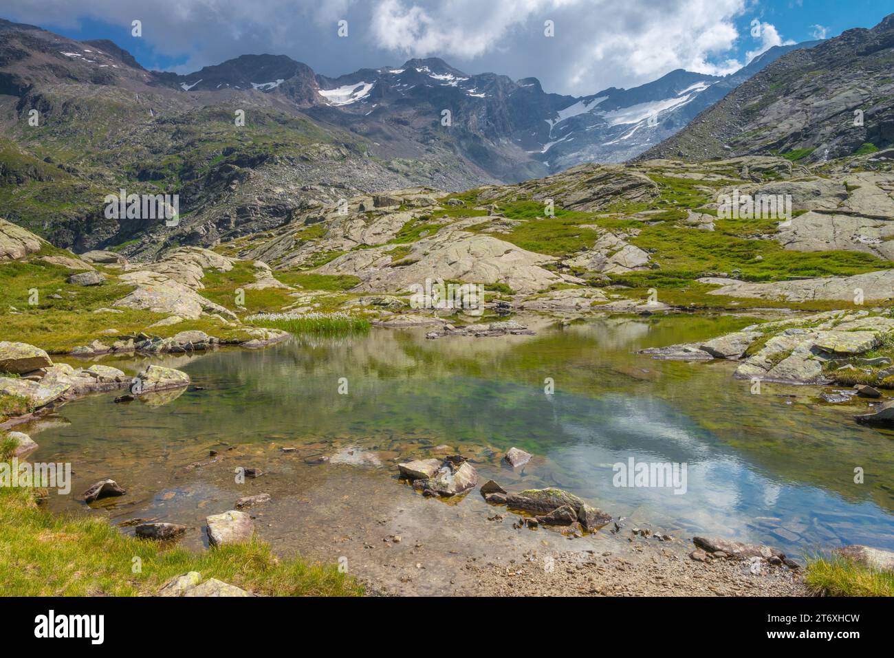 Majestic mountain scene in the Italian Alps, imposing mountain range with small glacier towering over valley with small alpine pond and rock slabs. Stock Photo