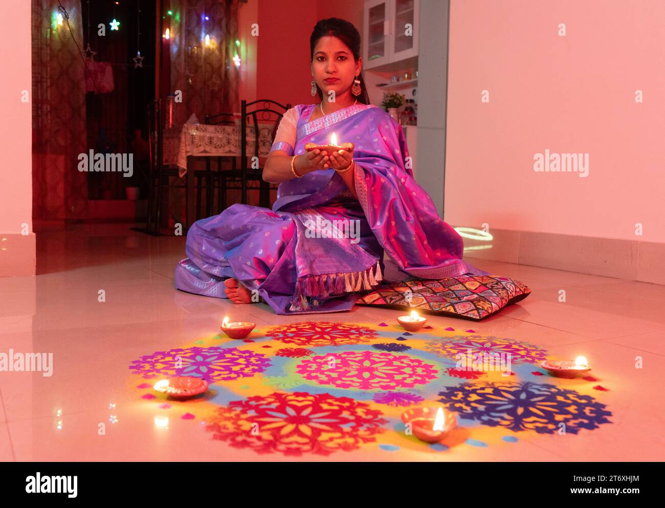 A woman holds an oil lamp sitting next to a rongoli, wearing