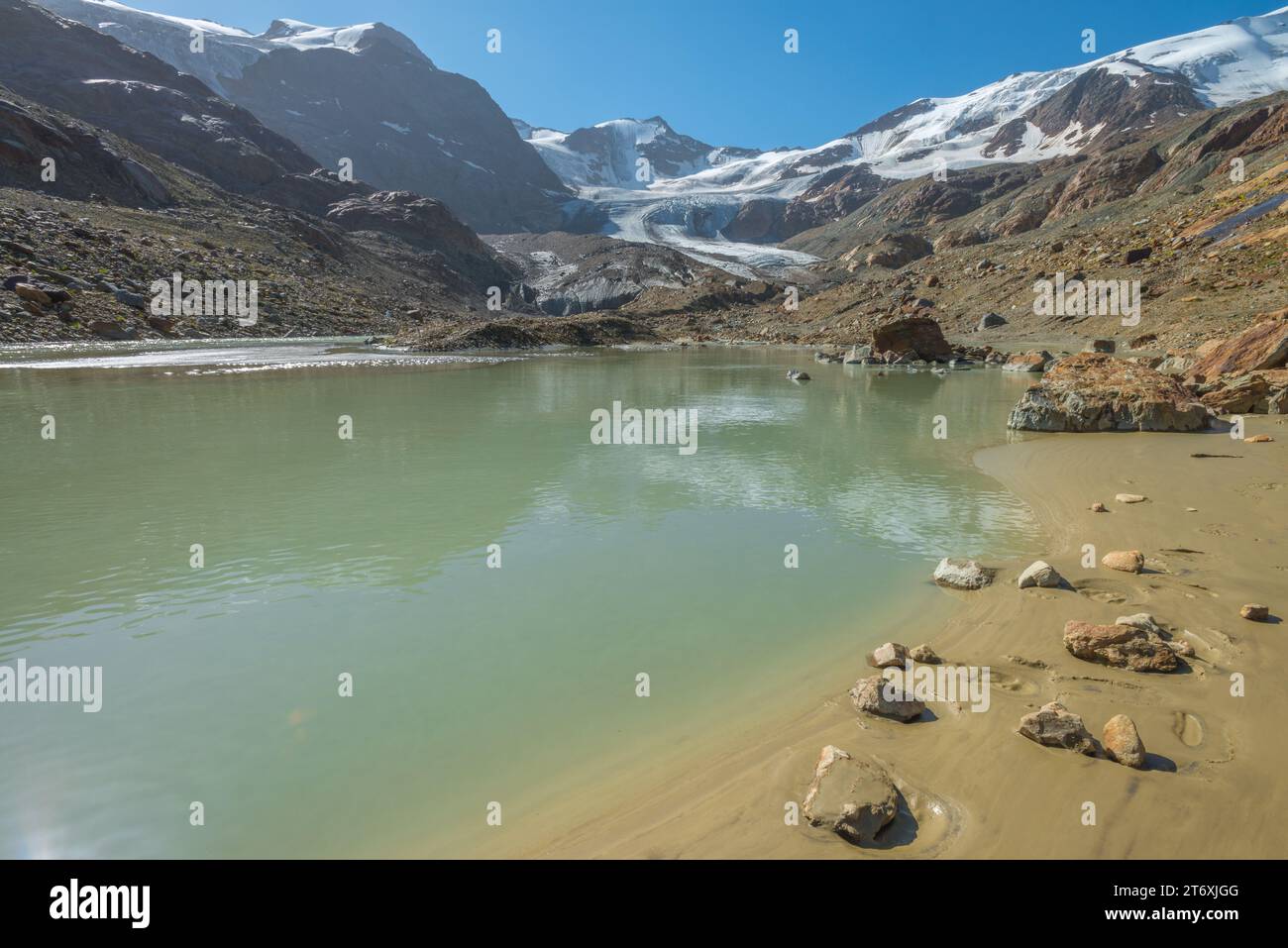 Forni Glacier in the Italian Alps, with glacial tongue melting onto a glacial stream loaded with silt. Small beach with wet sand and stones. Stock Photo