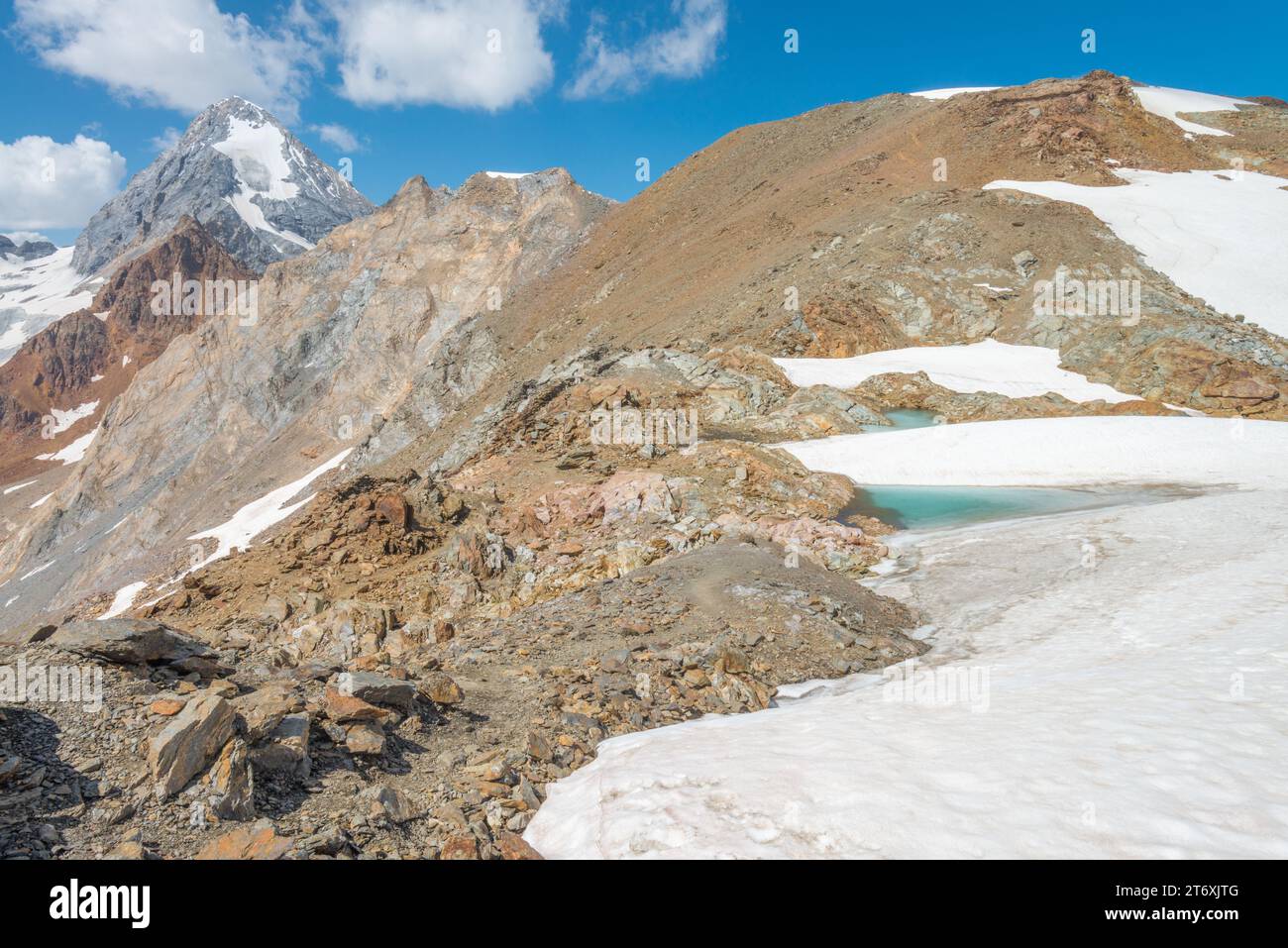 Hiking a dark mountain ridge, with small snowfield and meltwater tarn. Majestic prominent peak Konigspitze and glacier in the background Stock Photo