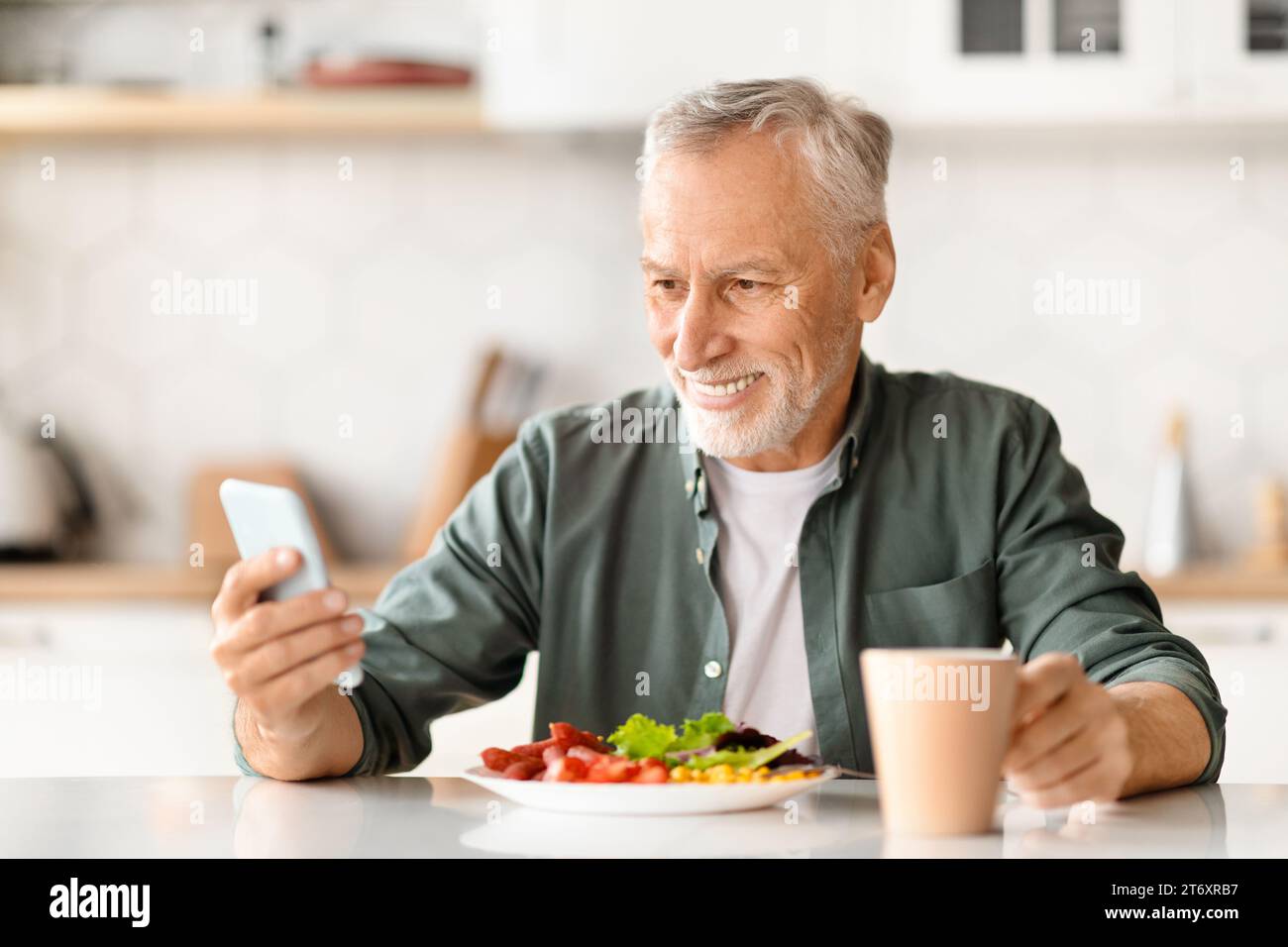 Happy senior man using smartphone while having breakfast at kitchen Stock Photo
