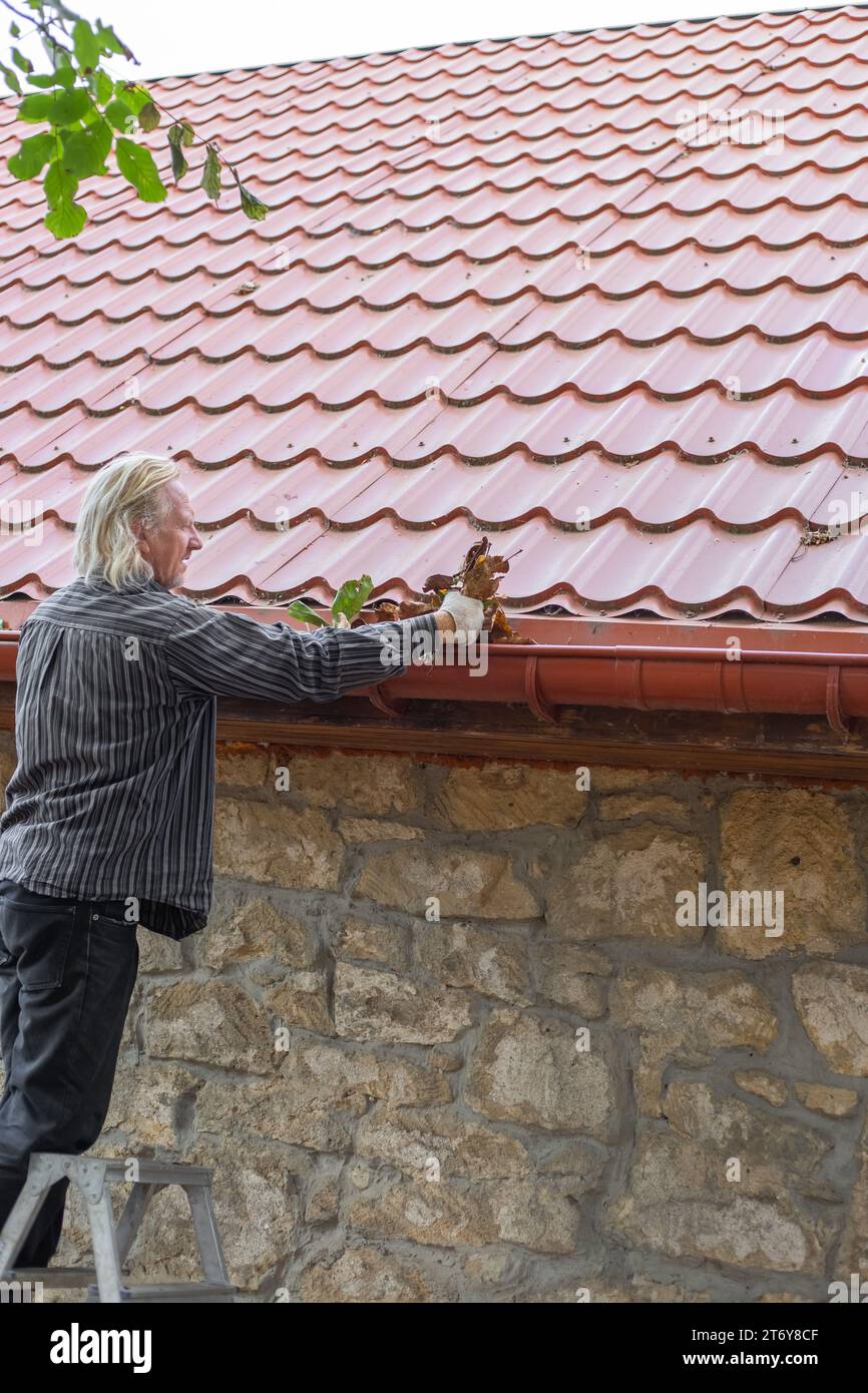 A mature man removes leaves and debris from the gutter of his house ...