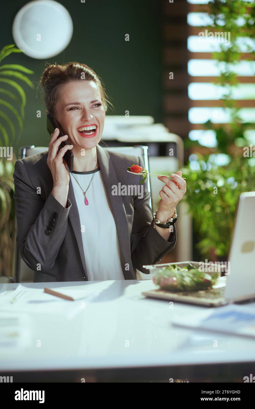 Sustainable workplace. smiling modern accountant woman in a grey business suit in modern green office with laptop eating salad and talking on a smartp Stock Photo