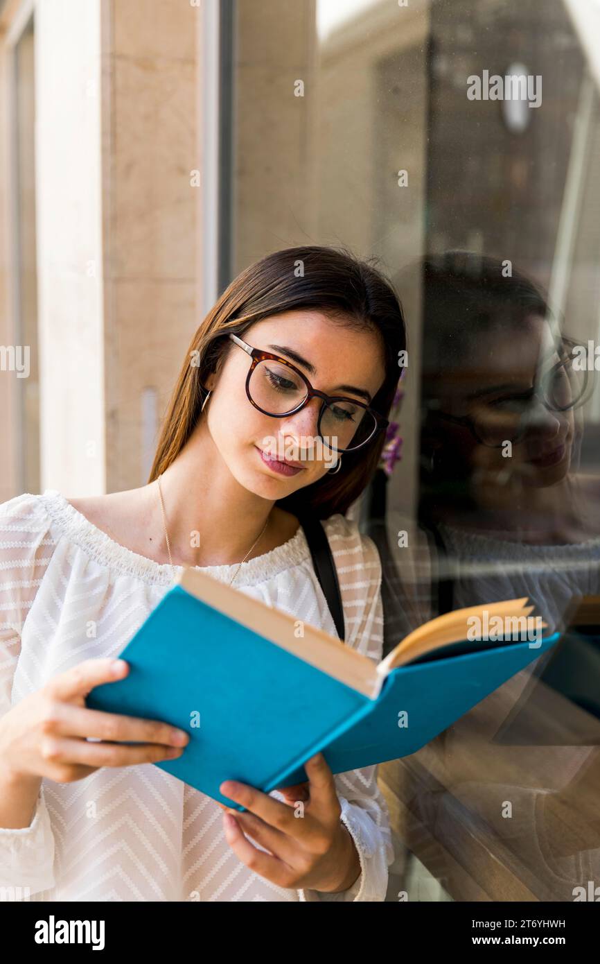 Young lady with glasses reading book near window Stock Photo