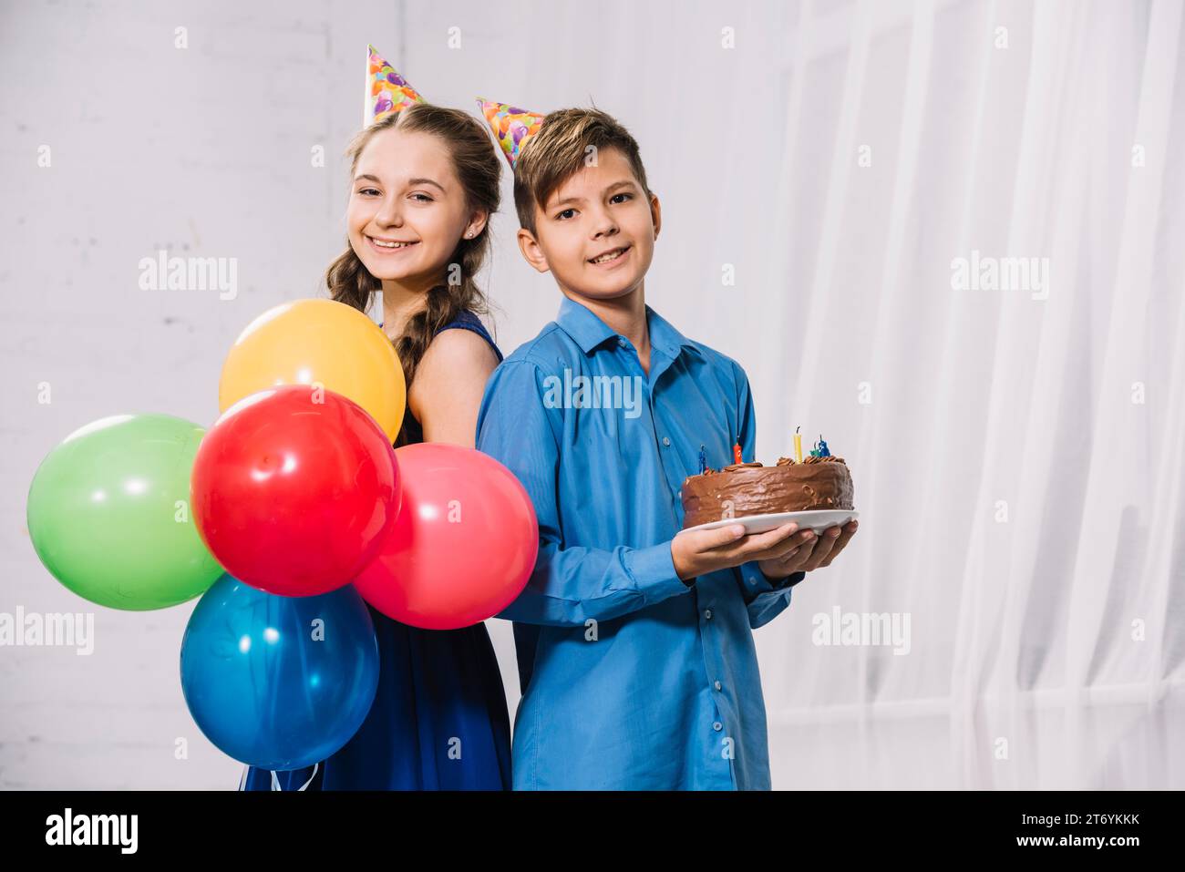 Girl holding balloons boy holding birthday cake standing back back looking camera Stock Photo